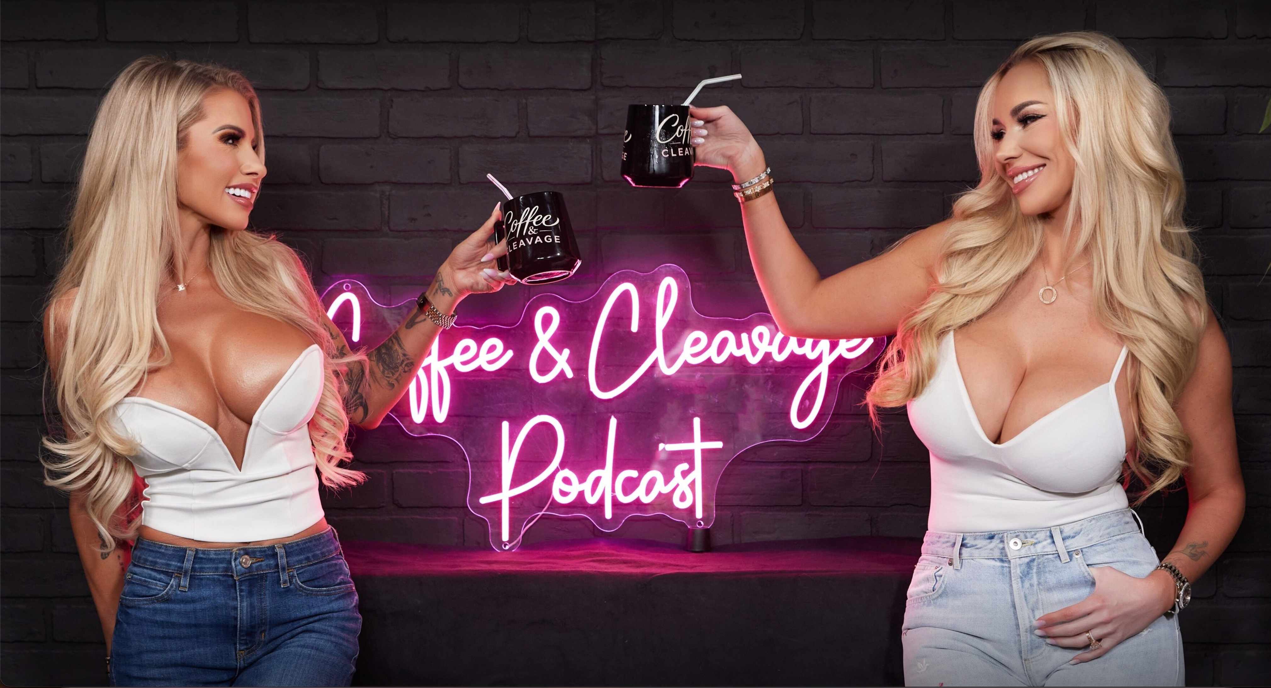 Two Coffee and Cleavage podcast hosts raising black coffee mugs in front of a pink neon show sign